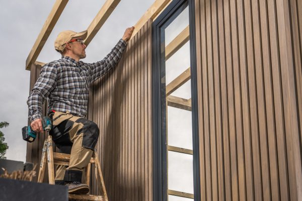 Experienced Caucasian Woodworker with Cordless Drill Driver in His Hands Building Modern Backyard Garden Shed. Composite Material Walls.